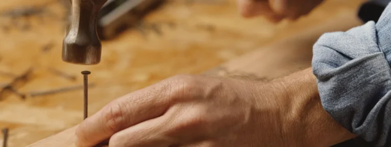 Nahaufnahme einer Hand die mit einem Hammer einen Nagel in hellbraunes Holz.