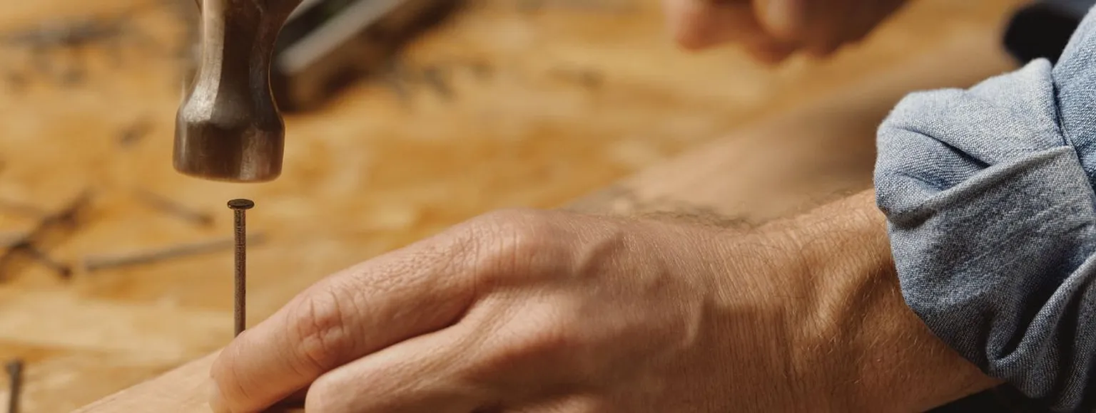 Nahaufnahme einer Hand, die mit einem Hammer einen Nagel in hellbraunes Holz auf einer Werkbank schlägt.