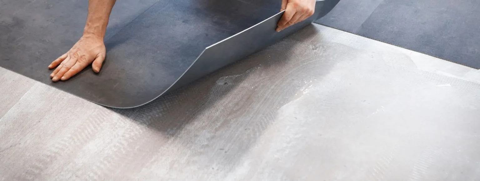 Nahaufnahme von Händen, die einen hellbraunen Vinyl-Bodenbelag auf einer glatten, hellen Oberfläche in einem Innenraum verlegen.