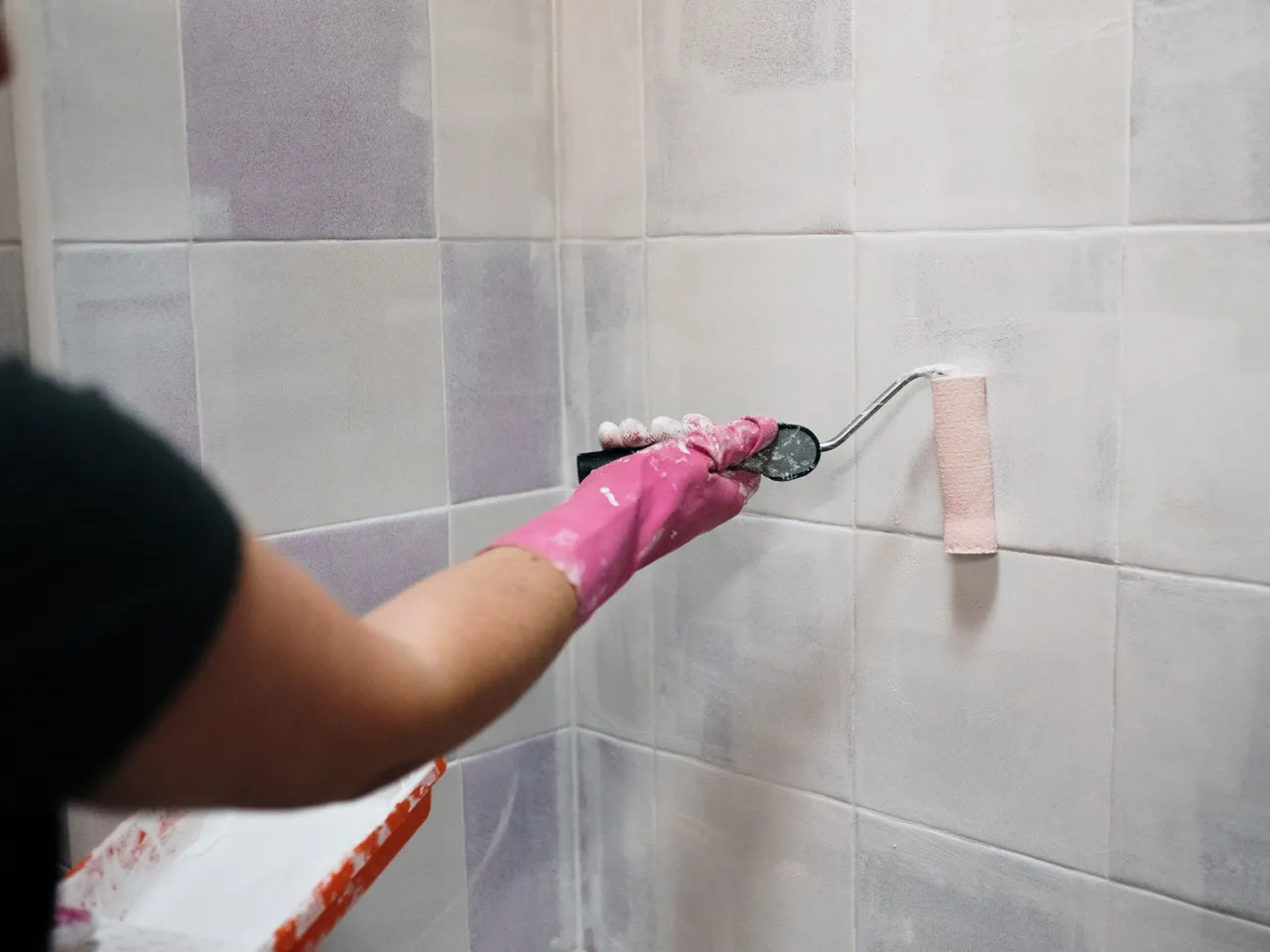 Person bemalt Badezimmerfliesen mit weißer Farbe, trägt pinken Handschuh beim Streichen.