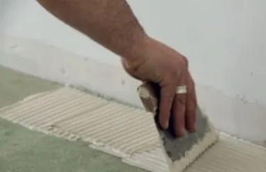 Person, die den hellgrauen Fliesenkleber vor dem Verlegen der Platten mit einer Zahnspachtel auf den Boden aufträgt.