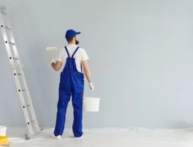 Person in blauen Overalls streicht im Winter mit Farbroller neben einem Leiter Innenraum.