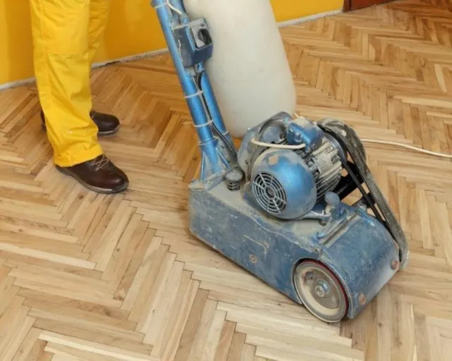 Person schleift helles Holzparkett mit einer Bodenschleifmaschine im Innenraum.