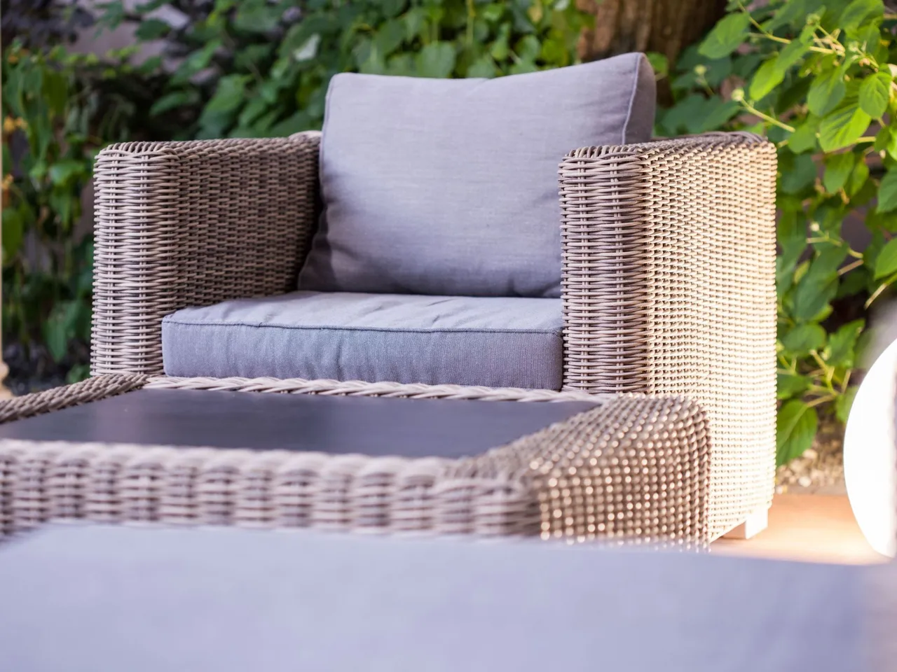 Terrassenstuhl aus Polyrattan mit grauen Kissen auf einer Terrasse, im Hintergrund ist Grünzeug zu sehen.