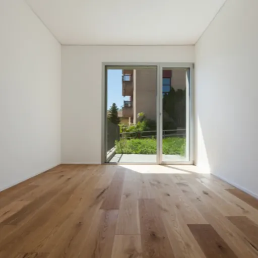Unmöbliertes Zimmer mit Holzparkettboden, weißen Wänden und Fenster mit Blick ins Grüne.