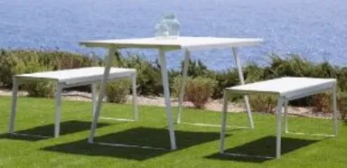 Weiße Metall-Gartenmöbel mit Tisch und Bänken im Gras am Meer, zwei Flaschen sichtbar.