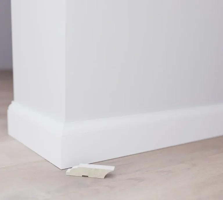 Weiße Sockelleiste, die mit einem kleinen Keil auf einem hellen Holzboden in einem Innenraum verlegt wird.