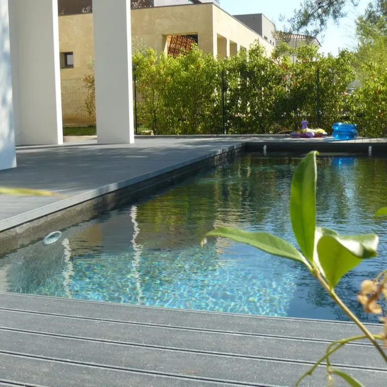 WPC-Pooldeck mit hellen Dielen, Grünpflanzen und einem Haus im Hintergrund.