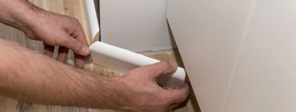 Hände montieren weiße Sockelleiste an hellem Holzfußboden