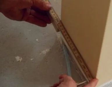 Zwei Hände messen mit einem gelben Zollstock eine weiße Wandecke für den Türeinbau aus.