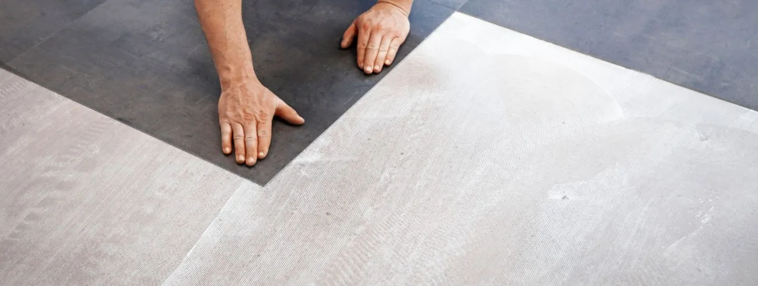 Zwei Hände verlegen dunkle Vinylbodenpaneele auf heller Fläche mit sichtbarer Fußbodenheizung.