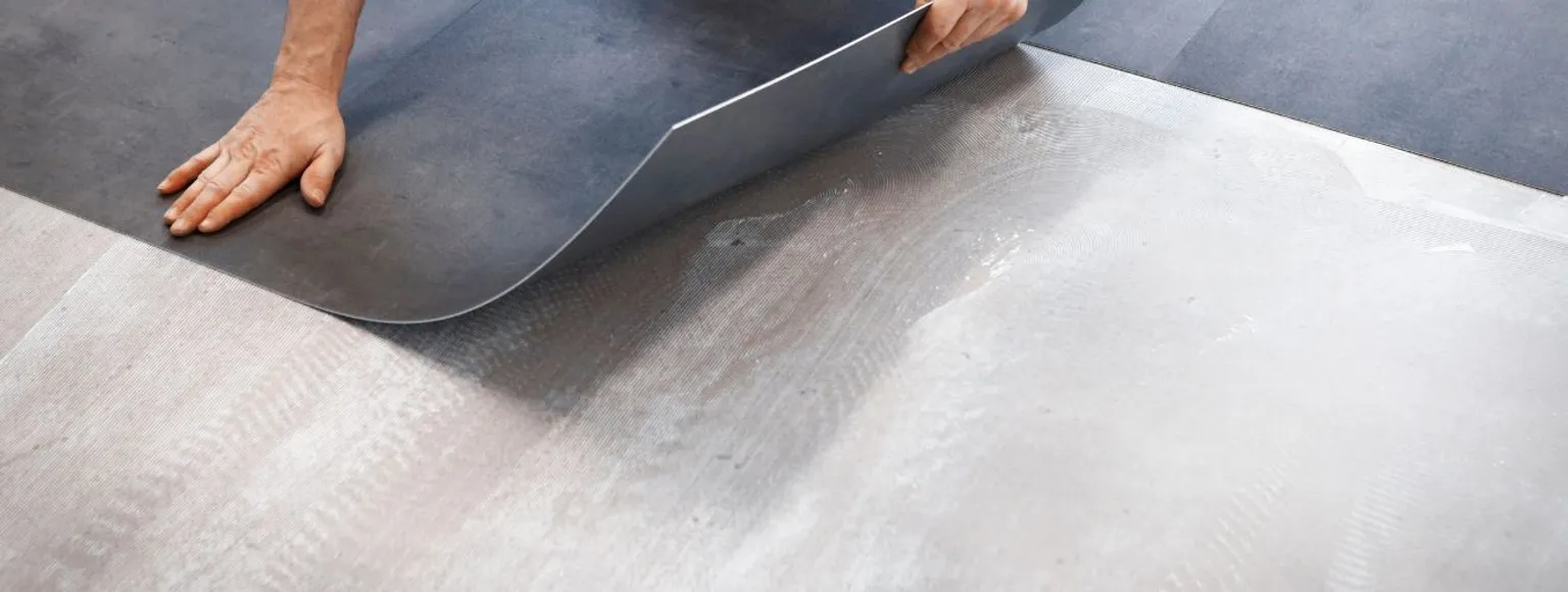 Zwei Hände verlegen hellbraune Vinyl-Bodenbelagsdielen auf einem schlichten grauen Unterboden in einem Innenraum.