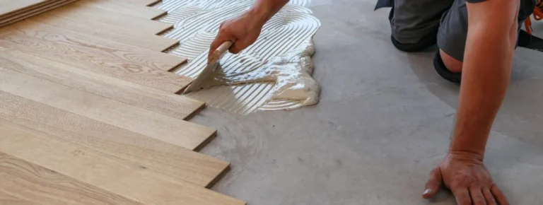 Hände verteilen Klebstoff auf Betonboden und verlegen helle Holzdielen