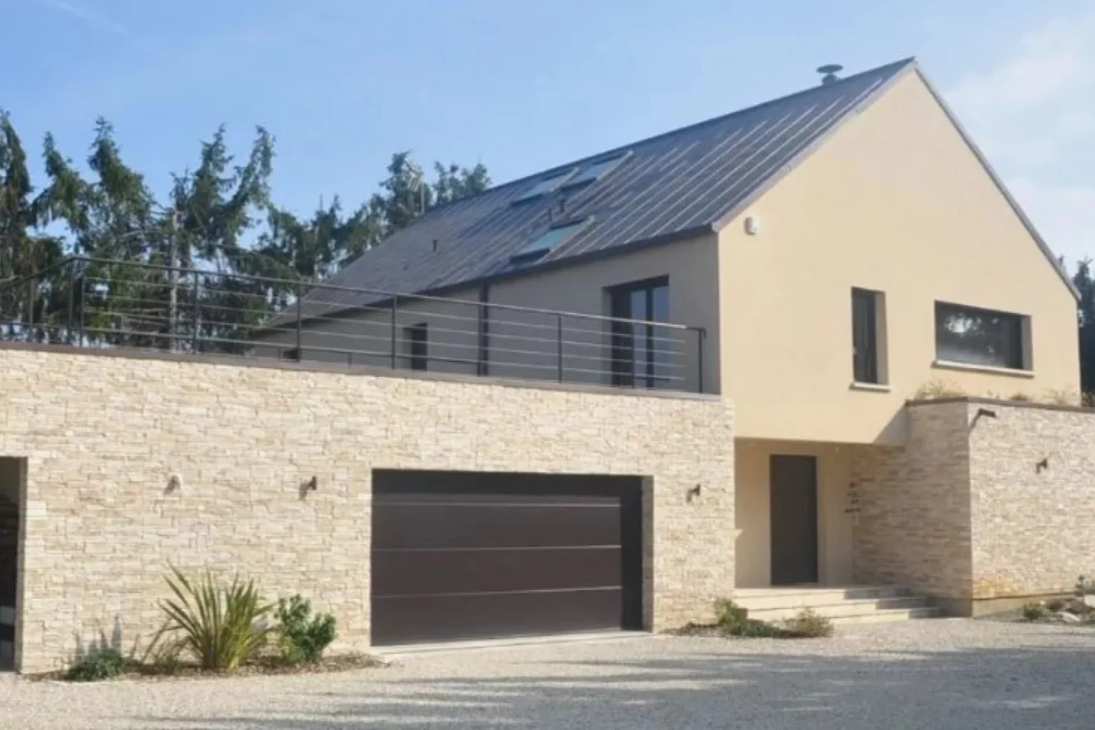 Das Bild zeigt ein modernes zweistöckiges Haus mit einer beigen Fassade und einem dunklen Dach. Der zweite Stock verfügt über einen großen Balkon, der mit einem Metallgeländer ausgestattet ist. Eine breite Auffahrt führt zu einem dunklen Garagentor, das die minimalistische Gestaltung des Eingangsbereichs ergänzt. In der Umgebung des Eingangs befinden sich nur wenige Pflanzen, was auf eine zurückhaltende Gartengestaltung hindeutet. Die schlichte Ästhetik des Hauses wird durch klare Linien und dezente Farben betont.