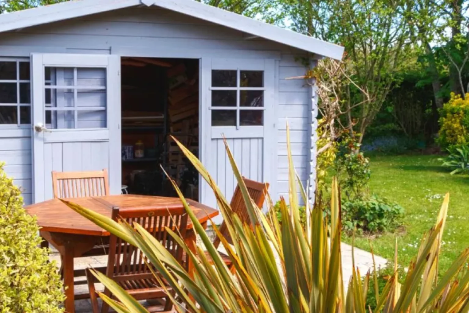 Ein hellblaues hölzernes Gartenhaus steht im Mittelpunkt des Bildes. Die doppelten Türen und Fenster des Schuppens sind geöffnet, was eine einladende Atmosphäre schafft. Vor dem Gartenhaus befindet sich ein Holztisch mit passenden Stühlen, die zum Verweilen und Entspannen einladen. Das gesamte Arrangement ist von einer Vielzahl grüner Pflanzen und Büsche umgeben, die den saftigen Rasen ergänzen und für eine ruhige Gartenszene sorgen. Diese harmonische Umgebung bietet einen idealen Rückzugsort im Freien, um sich zu erholen oder gesellige Stunden zu verbringen.