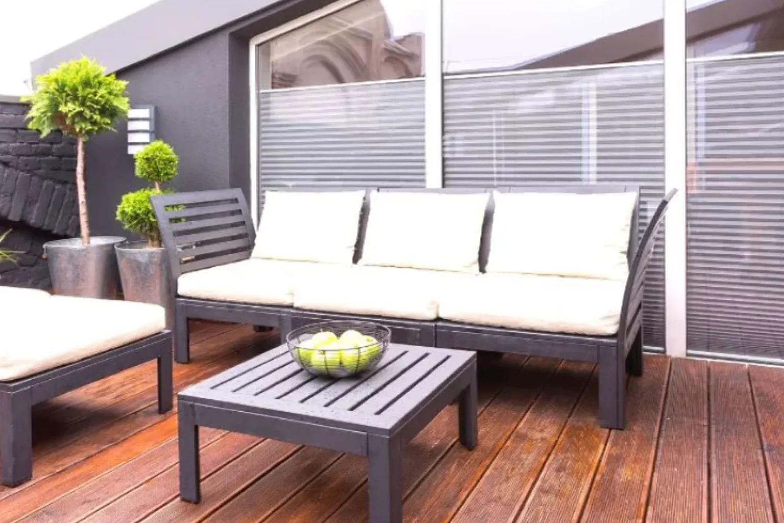 Das Bild zeigt eine Outdoor-Patio, die nahtlos in den angrenzenden Garten übergeht. Im Vordergrund steht ein modernes Sofa aus schwarzem Metall mit weißen Sitz- und Rückenpolstern. Vor dem Sofa ist ein passender Couchtisch in derselben schwarzen Metalloptik platziert. Auf dem Tisch befindet sich eine Schale mit grünen Äpfeln als Dekorationselement. Der Boden der Terrasse ist mit Holzplanken gestaltet, was für eine natürliche Atmosphäre sorgt. Um den Bereich herum sind verschiedene Topfpflanzen aufgestellt, die zur grünen Umgebung des Gartens harmonisch passen und der Balkon-Oase einen zusätzlichen Hauch von Eleganz verleihen.