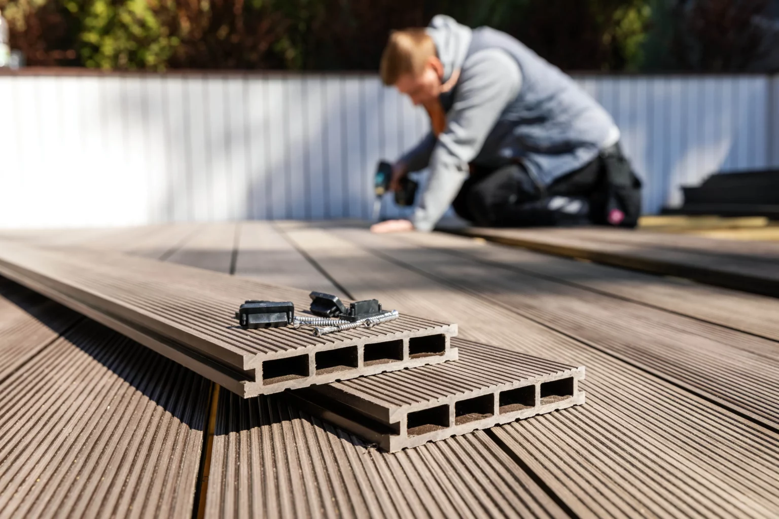 Eine Person hockt auf ein Holzdeck im Garten und verwendet ein Werkzeug, das leicht unscharf ist. Im Vordergrund sind zwei Stapel mit Verbundholz-Dielen erkennbar, auf denen Schrauben und Befestigungsclips liegen. Der Hintergrund besteht aus einem Zaun und üppigem Grün. Die Szene vermittelt den Eindruck von Gartenarbeit oder Heimwerkeraktivitäten im Freien, wobei der Fokus auf der Installation oder Wartung von Terrassendielen liegt. Die natürliche Umgebung schafft eine ruhige und produktive Atmosphäre für die Arbeit im Freien.