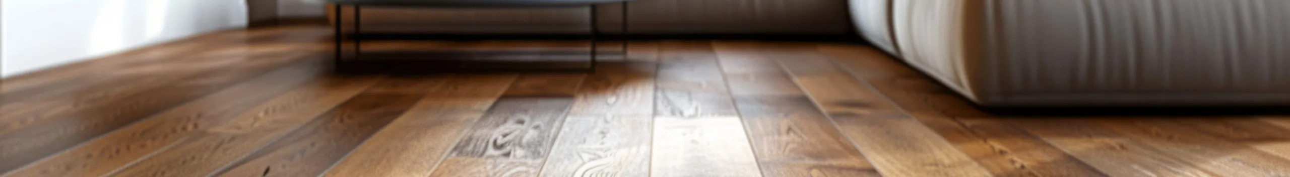 Nahaufnahme eines mitteltönigen Holzbodens, im Hintergrund sind Sofa und Tisch zu sehen.