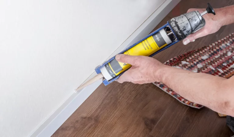 Eine Person verwendet eine weiße Kartuschenpistole, um weißen Dichtstoff entlang des Sockels einer weißen Wand aufzutragen. Der Boden besteht aus braunem Holzlaminat und bietet einen warmen Farbkontrast zur Wand. Auf der rechten Seite des Bildes ist teilweise ein Teppich mit rotem Muster zu sehen. Die Szene vermittelt den Eindruck einer präzisen Arbeit, bei der die Person sorgfältig darauf achtet, eine gleichmäßige Abdichtung herzustellen.