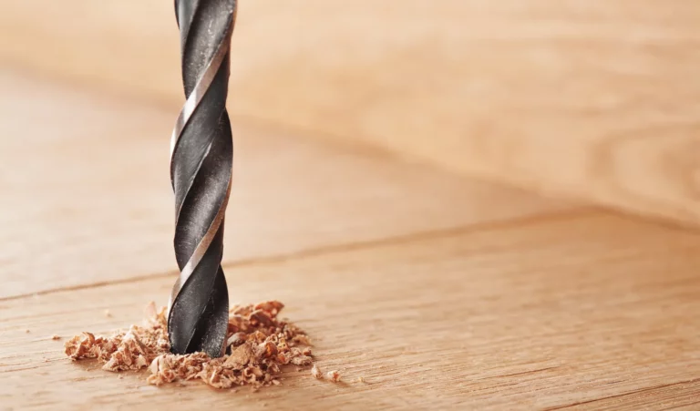 Nahaufnahme eines Bohrers, der in eine hölzerne Oberfläche eindringt. Der Bohrer ist silbern und durchdringt das helle, glatte Holz schnell vollständig. Rund um den Bohrer sind kleine Holzspäne zu sehen, die durch den Bohrauftrag entstanden sind. Der Hintergrund ist verschwommen und in einem sanften Braunton gehalten, wodurch der Fokus auf dem handwerklichen Arbeitsprozess liegt. Die Szene vermittelt einen Eindruck von Heimwerken oder handwerklicher Tätigkeit im Bereich Haus und Garten.