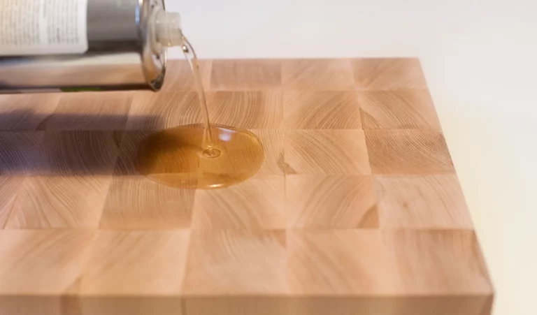 Ein klarer Alt-Text für das beschriebene Bild könnte folgendermaßen gestaltet werden: „Eine Flasche gießt Öl auf ein hölzernes Schneidebrett. Das Schneidebrett hat ein kariertes Muster, bei dem die Holzmaserung deutlich erkennbar ist. Auf der Oberfläche des Brettes bildet sich eine kleine Ölpfütze, was darauf hindeutet, dass es zur Pflege oder Wartung vorbereitet wird.“.
