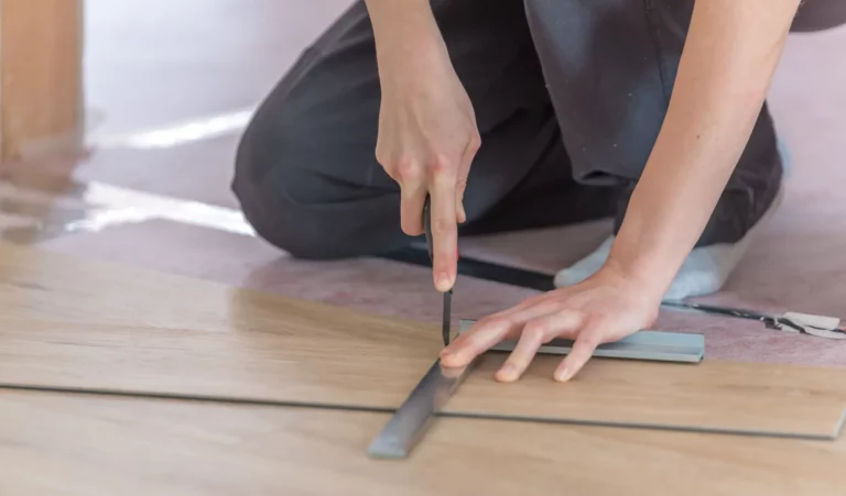 Ein Mensch kniet auf einem Holzboden und schneidet mit einem Universalmesser ein Stück Holz. Eine Metallleiste dient als Schneideführung, um präzise Linien zu gewährleisten. Die Hände der Person sind das zentrale Element des Bildes, während ein Teil ihrer Beine im Hintergrund sichtbar ist. Der Boden, auf dem gearbeitet wird, hat eine natürliche Holzmaserung und vermittelt einen warmen Ton. Um die Person herum sind keine weiteren Gegenstände oder Personen zu sehen, war der Fokus klar auf die handwerkliche Tätigkeit gerichtet. Die neutrale Beleuchtung sorgt für eine klare Sicht auf das Arbeitsmaterial und das Werkzeug.