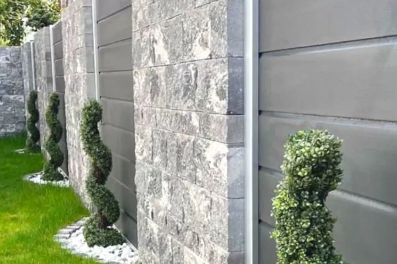 Das Bild zeigt eine Reihe von spiralförmigen Formschnitt-Gehölzen, die im Garten entlang eines Zauns aus Stein und Metall angeordnet sind. Die Heckenpflanzen wirken gepflegt und kunstvoll geschnitten, was auf eine sorgfältige Gartenpflege hingewiesen wird. Unter den Pflanzen sind kleine weiße Steine zu sehen, die einen optischen Kontrast zu dem umgebenden saftig grünen Gras bilden. Diese weiße Steinfläche grenzt sich klar vom restlichen Rasen ab und verleiht der Gartengestaltung eine strukturierte Ästhetik. Der Gesamteindruck ist ein harmonischer Mix aus natürlicher Schönheit und geordneter Gestaltung, der sowohl praktische als auch dekorative Zwecke erfüllt. Es gibt keine Personen oder Tiere auf dem Bild; Der Fokus liegt vollständig auf den Pflanzen und der Anlage des Gartens sowie dem Wechselspiel mit den verwendeten Materialien wie Stein und Metall. Die Szene vermittelt ein Gefühl von Ruhe und sorgfältiger Planung in dieser grünen Oase.