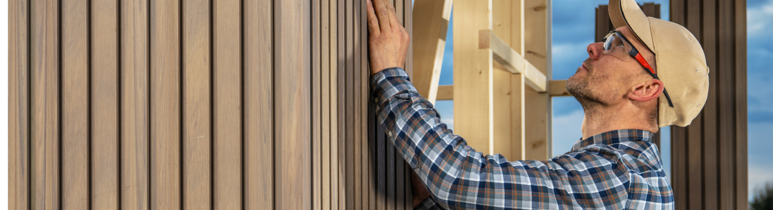 Ein Mann mit einer Mütze bringt hellbraune vertikale Holzverkleidungen an einer Hauswand im Freien an.