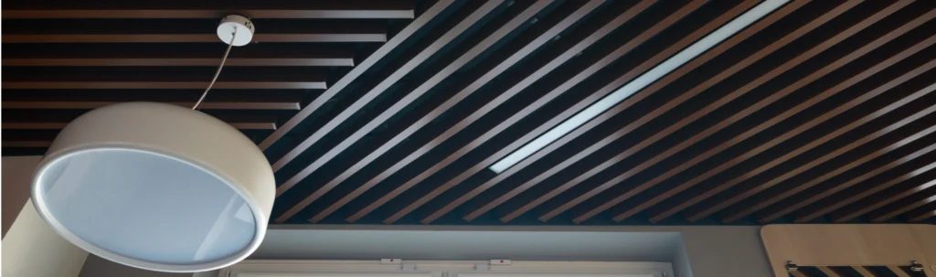 Detailaufnahme einer modernen Decke mit dekorativen, schräg verlaufenden Lamellenpaneelen in Holzoptik. Zwischen den Lamellen ist eine integrierte LED-Leuchte eingelassen, die für funktionale Beleuchtung sorgt. Die Kombination aus dunklem Holzdesign und technischer Ausstattung schafft eine stilvolle und akustisch optimierte Raumdecke.