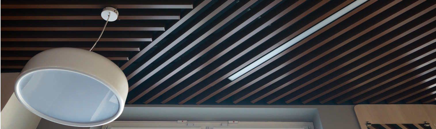 Detailaufnahme einer modernen Decke mit dekorativen, schräg verlaufenden Lamellenpaneelen in Holzoptik. Zwischen den Lamellen ist eine integrierte LED-Leuchte eingelassen, die für funktionale Beleuchtung sorgt. Die Kombination aus dunklem Holzdesign und technischer Ausstattung schafft eine stilvolle und akustisch optimierte Raumdecke.