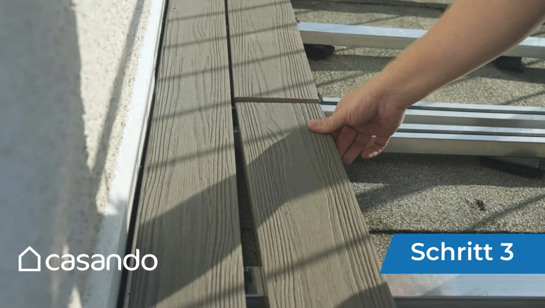 Eine Person legt während der Montagearbeiten Terrassendielen auf die Unterkonstruktion einer Terrasse.