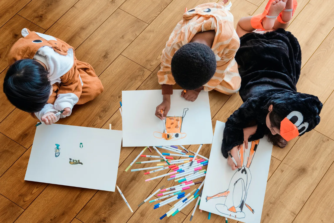 Drei Kinder zeichnen mit Papier und Stiften auf einem hellen Bio-Vinylboden.