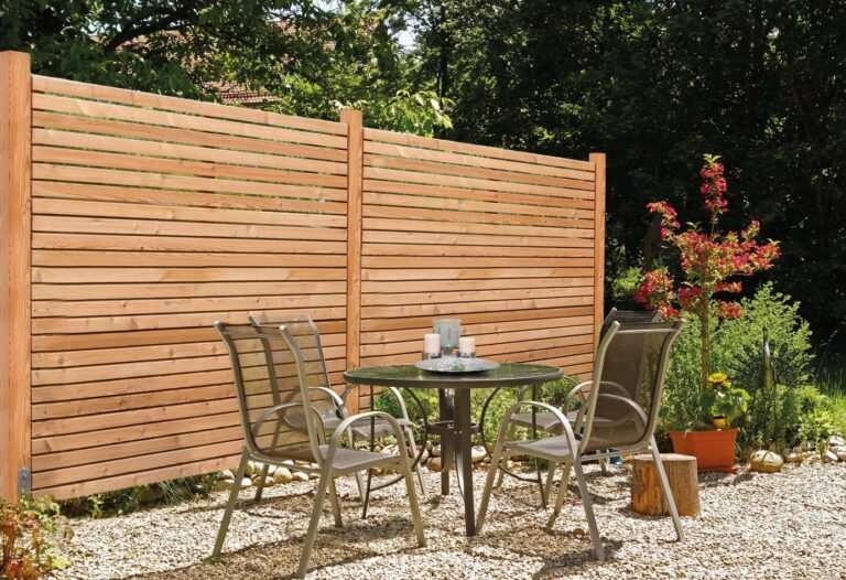 Rhombuszaun aus Holz mit horizontalen Leisten als Sichtschutz hinter einer Sitzgruppe in einem Garten.