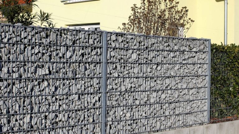 Gabionenzaun aus mit grauen Steinen als Sichtschutz vor einem Wohnhaus.