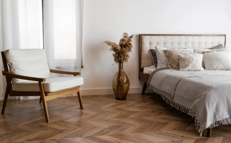 Modernes Schlafzimmer mit hochwertigem Fischgrät-Parkettboden, einem gemachten Bett mit vielen Kissen, einer Bodenvase mit Ziergräsern und einem Sessel am Fenster.