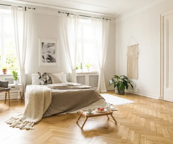 Ein lichtdurchflutetes Schlafzimmer mit edlem Fischgrät-Parkettboden, einem gemachten Bett, Deko-Pflanzen und einer Tasse Kaffee auf einem Betttablett auf dem Holzboden.