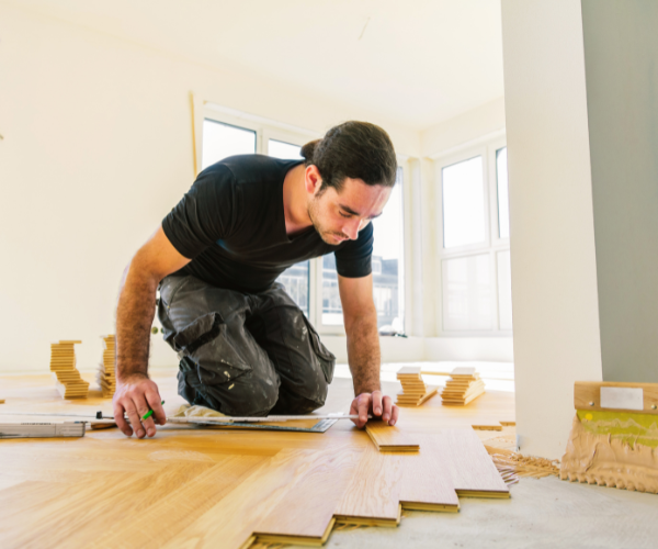 Ein Handwerker bzw. Bodenleger kniet auf dem Fußboden und misst beim Verlegen von Parkett präzise eine Holzdiele mit Werkzeug aus.
