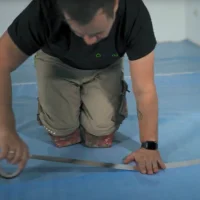 Eine Person kniet auf einem grauen Boden und klebt Nähte aus Hartvinyl mit Schallschutzschicht ab.