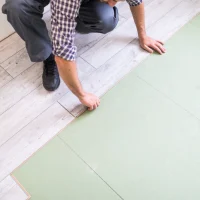 worker_processing_floor_with_bright_laminated_flooring_boards_1_1x Eine Person verlegt eine Trittschalldämmung unter einem Vinyl-Bodenbelag auf einer hellen Holzoberfläche in einem Innenraum.